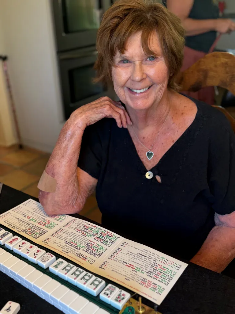 Nancy Guthrie, Savannah Guthrie's mother, seated at a table with mahjong tiles and a game card.