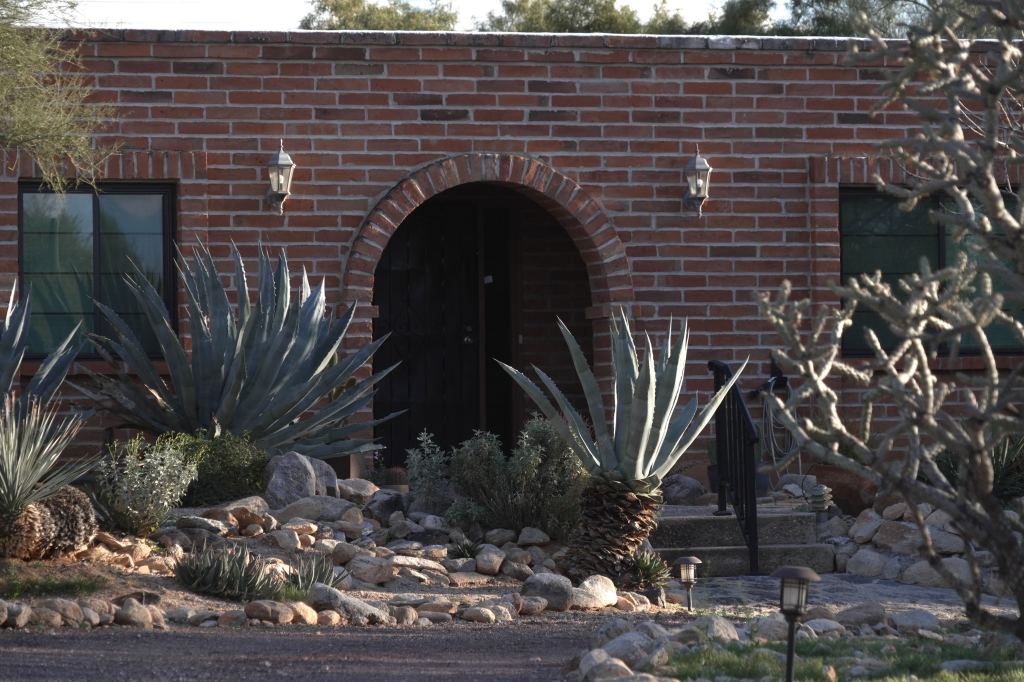The exterior of Nancy Guthrie's home in Tucson, Arizona, showing a brick wall, an arched doorway, and desert landscaping.