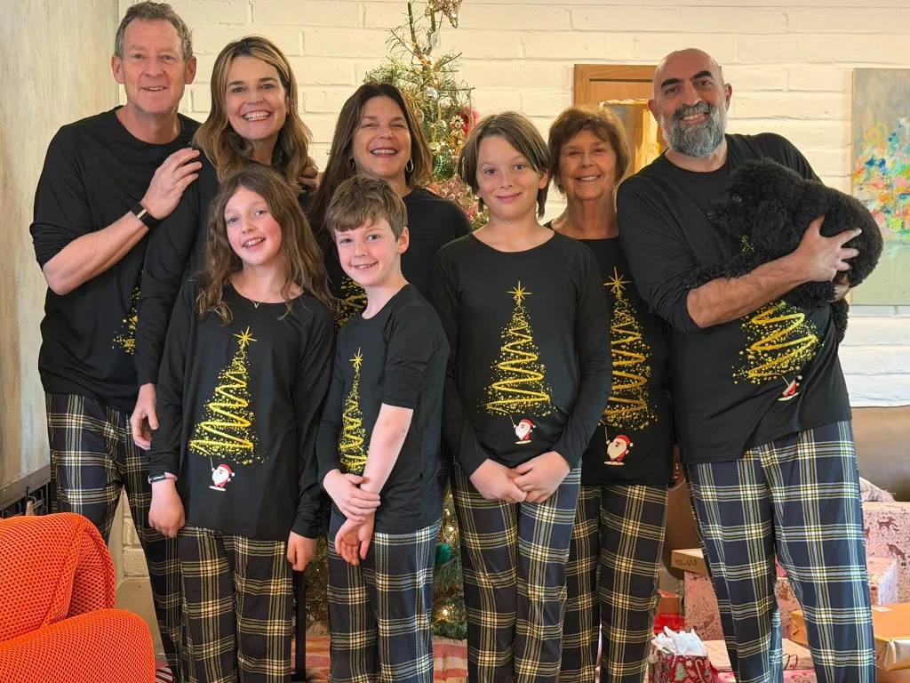 A family of eight, including a young girl, two boys, two adult women, an adult man, an elderly woman, and a man holding a black poodle, wearing matching black Christmas tree pajamas and plaid pants, standing in front of a Christmas tree.