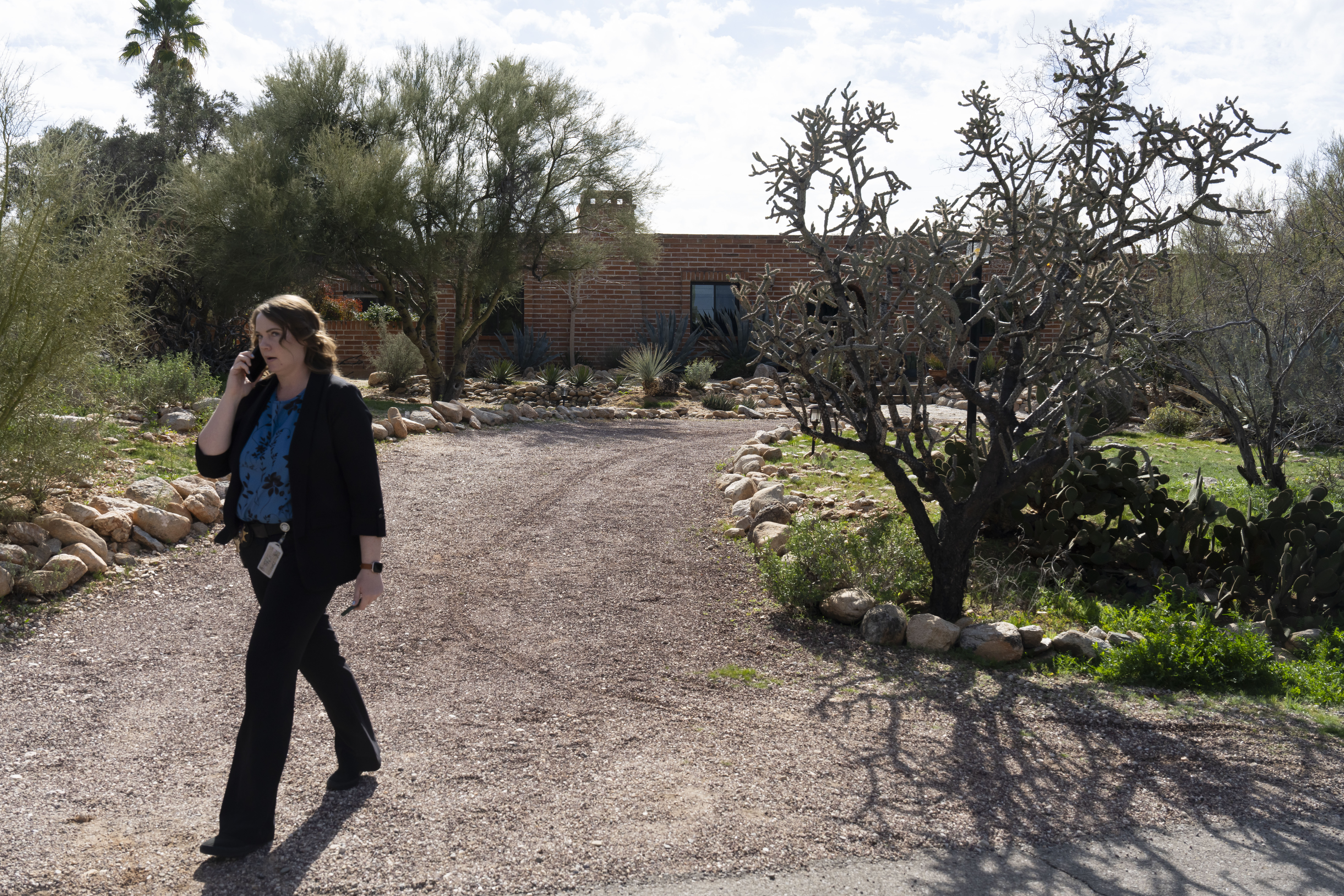 Woman walking on a gravel driveway in front of a brick house with desert landscaping, talking on a phone.