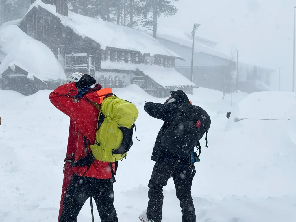 Two people in winter gear with backpacks stand in a snowy landscape near snow-covered buildings, likely searching for avalanche victims.