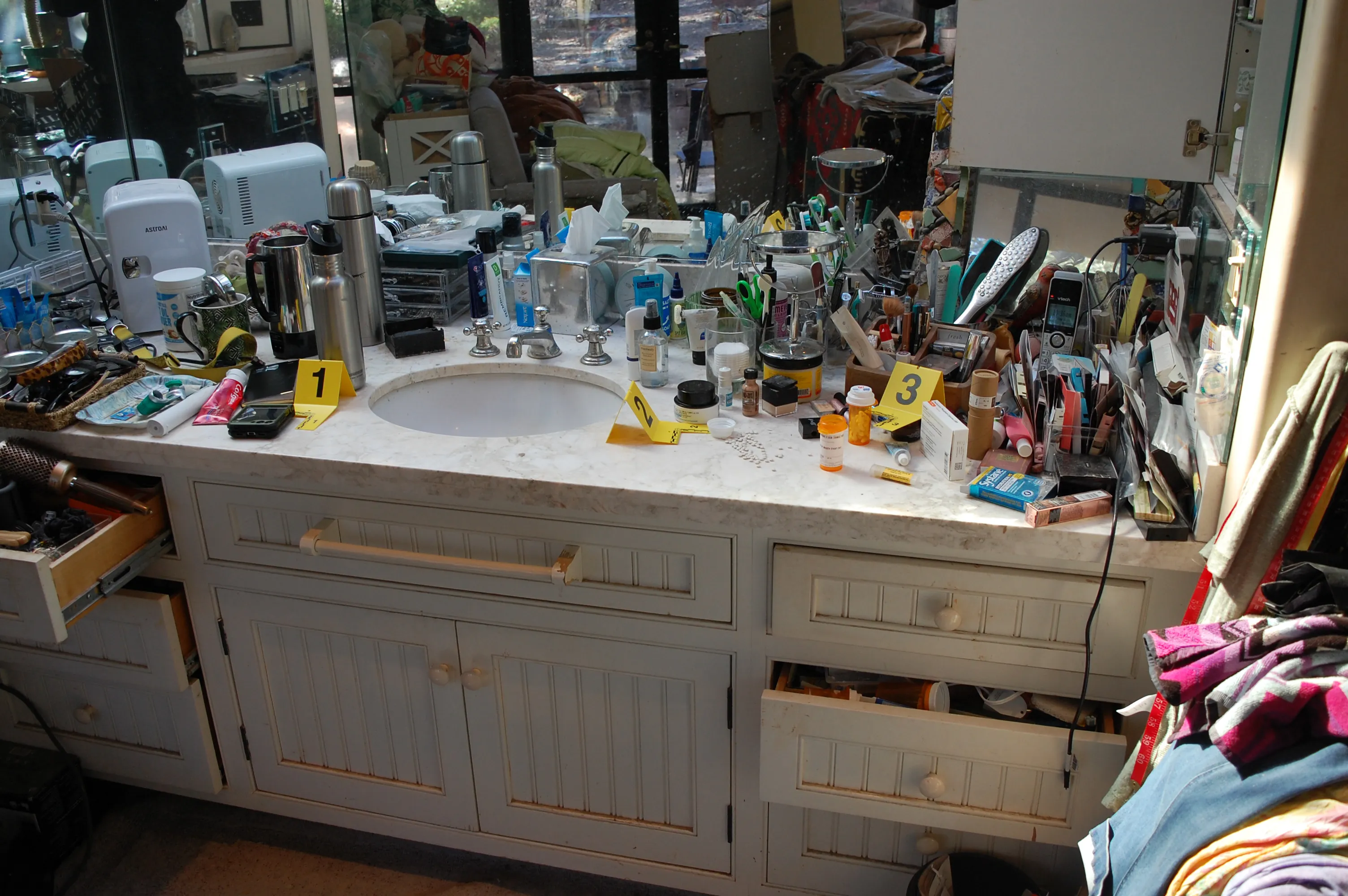 A messy bathroom vanity with a white marble countertop, featuring a sink surrounded by various toiletries and personal items, with some drawers ajar and items spilling out.