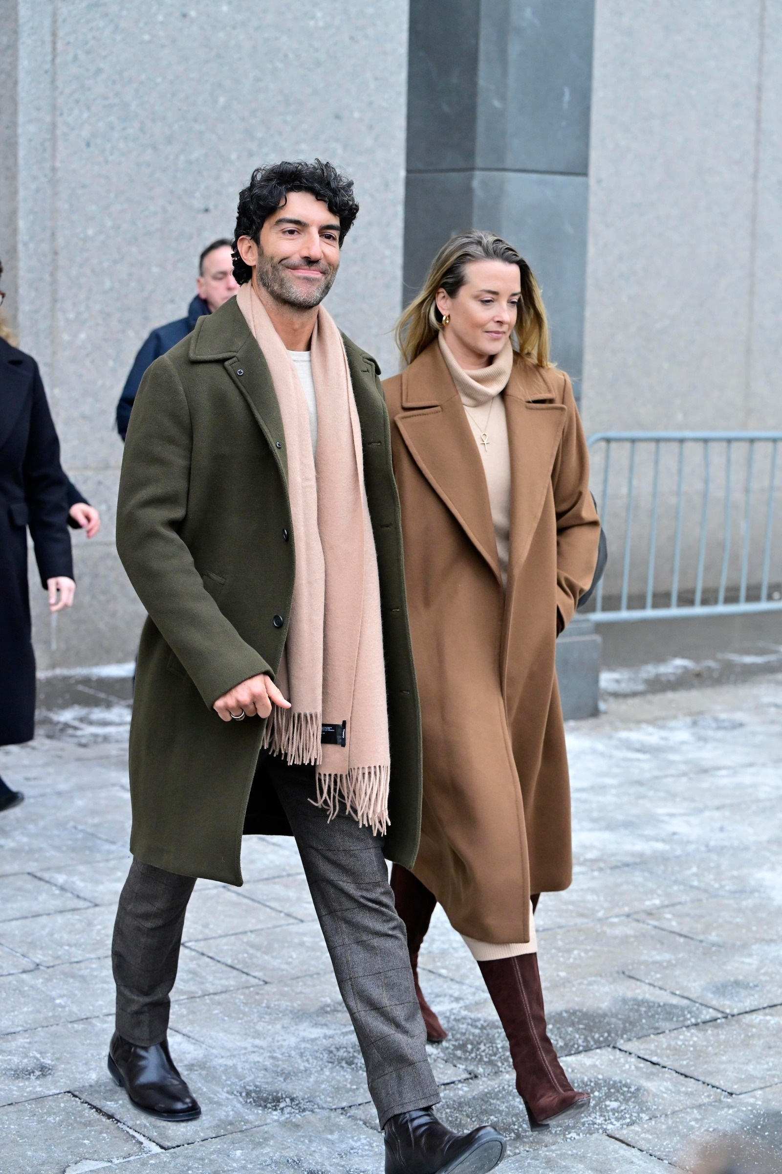 Justin Baldoni smiling and walking with a woman in a brown coat.