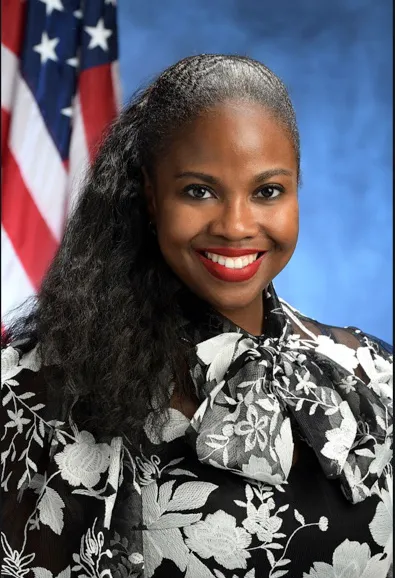 Headshot of New York State Assemblywoman Taylor Darling smiling in front of an American flag and a blue background.