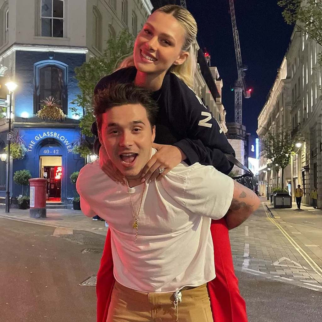 Nicola Peltz smiling while on Brooklyn Beckham's back, who is also smiling, on a street at night.