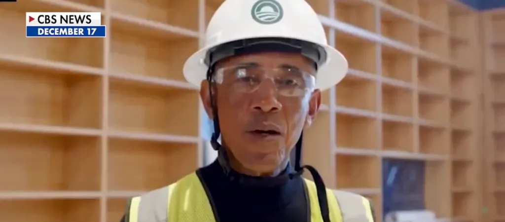 Barack Obama in a hard hat, safety glasses, and reflective vest, speaking indoors.