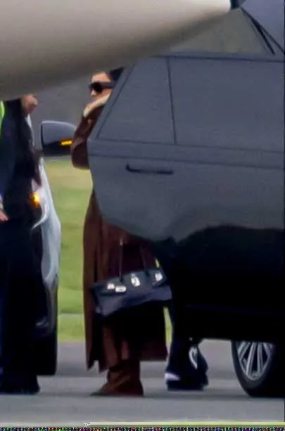 Kim Kardashian, wearing a brown coat and black sunglasses, holding a black Hermes Birkin bag, stands next to a car and a private jet.