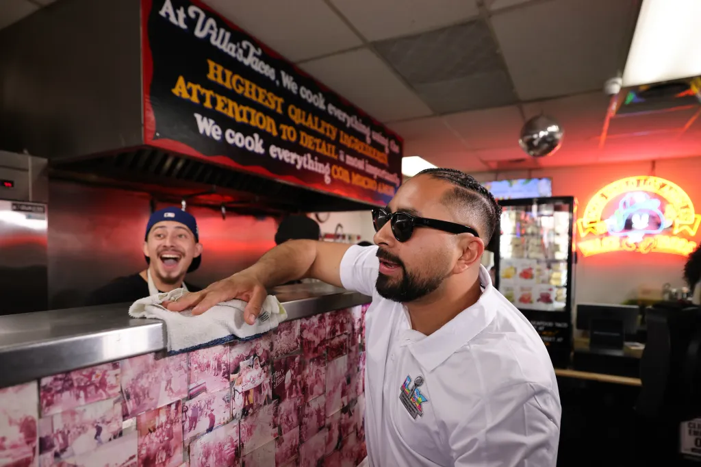 Owner Victor Villa, wearing sunglasses, speaks in front of a counter while another person cleans the counter in a taco restaurant.