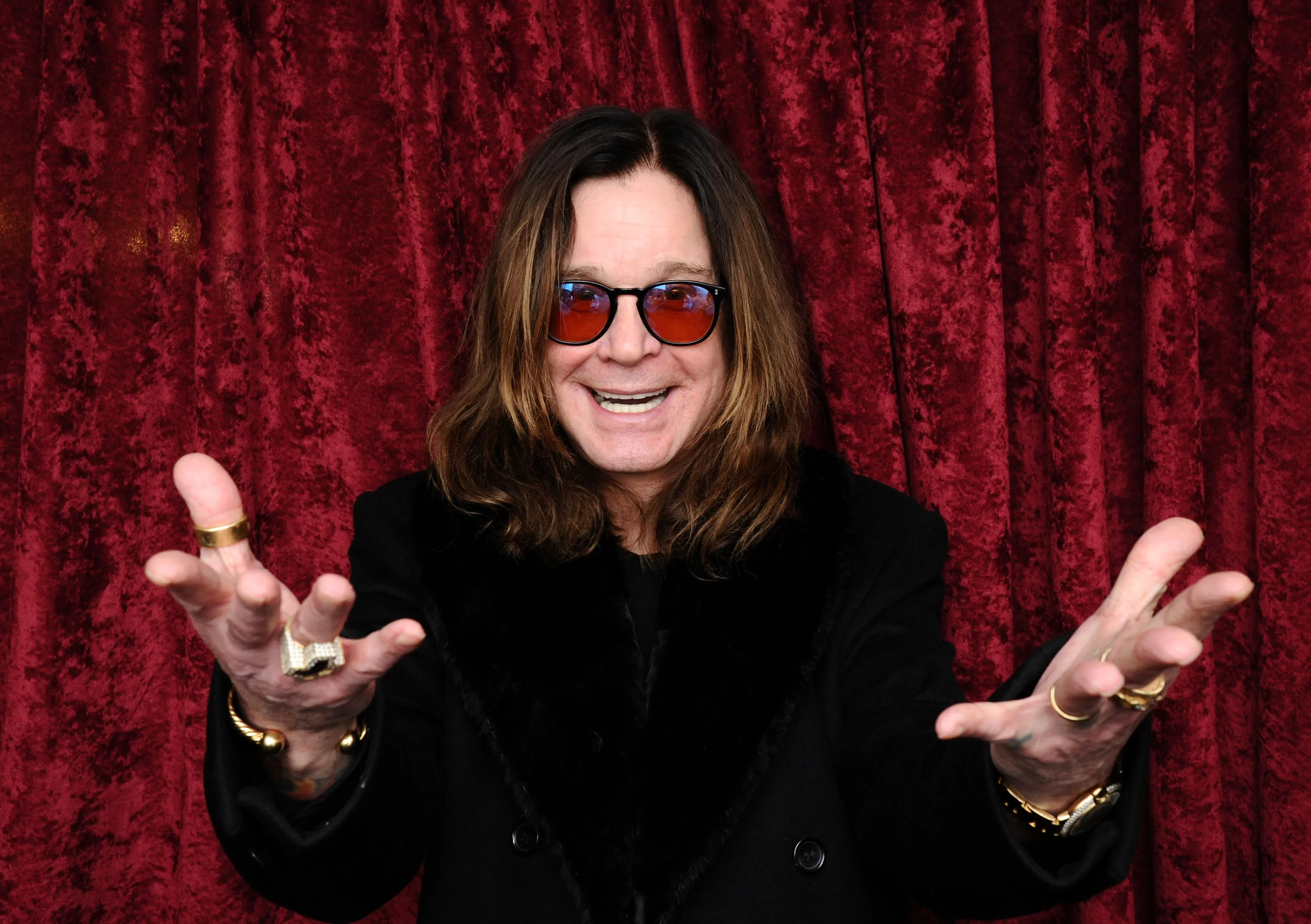 Ozzy Osbourne smiling with open hands, wearing red sunglasses and rings, against a red velvet background.