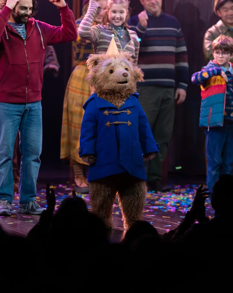 Paddington Bear, wearing a blue coat and party hat, stands center stage surrounded by actors and confetti, during the curtain call for 