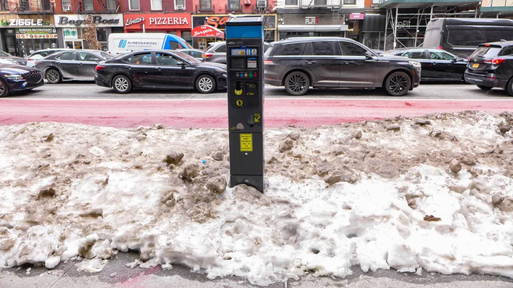 A parking meter on a New York City street is surrounded by plowed snow, with cars and storefronts in the background.