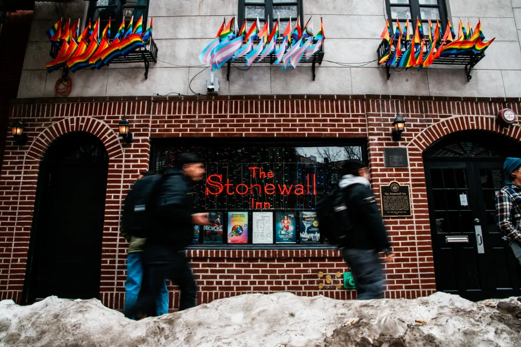 People walk past The Stonewall Inn with small rainbow and transgender pride flags displayed in window boxes above the entrance.