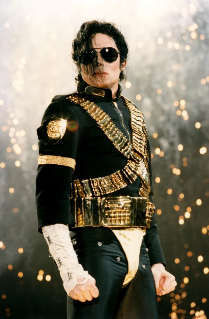 Michael Jackson performing on stage in a black jacket with gold accents and sunglasses, with hair falling over his face.
