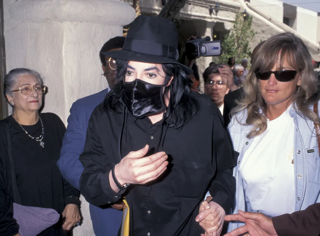 Michael Jackson, wearing a black hat and face mask, holding hands with Debbie Rowe, with an elderly woman beside them.