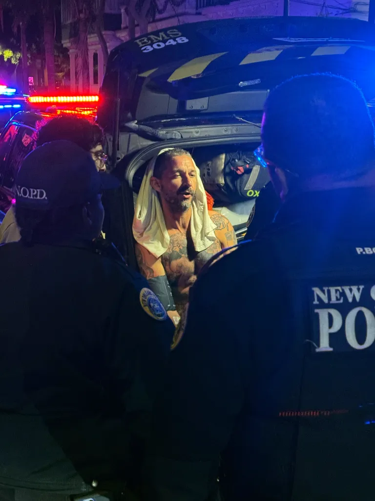 Actor Shia LaBeouf, shirtless and covered in tattoos with a white towel over his head, being apprehended by police at night.