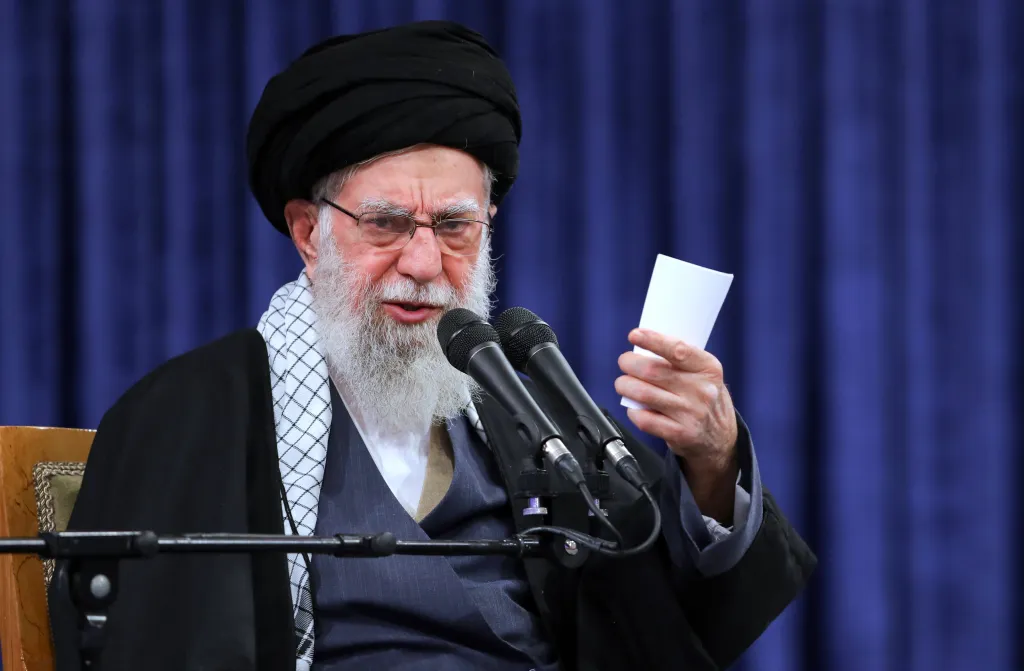 Supreme Leader Ayatollah Ali Khamenei speaking at a meeting in Tehran, holding a piece of paper.