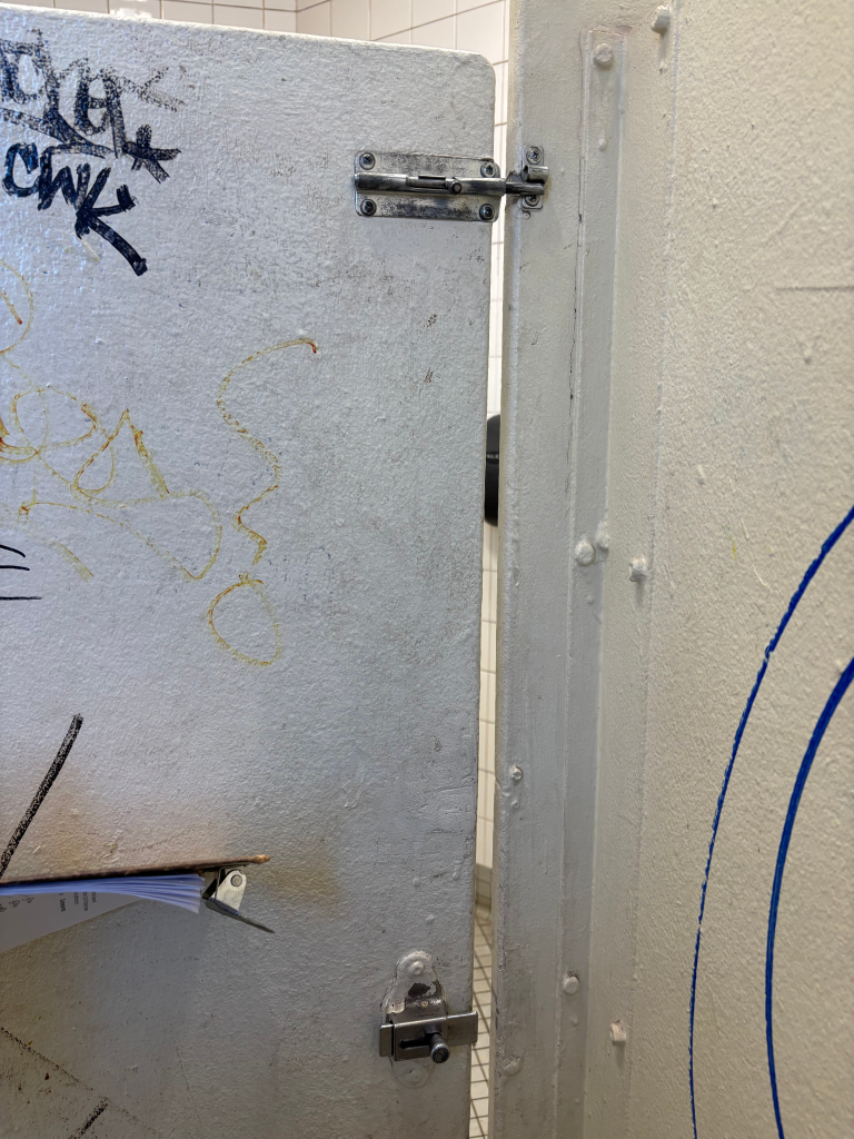 A grimy door with graffiti and a broken lock in a subway men's bathroom.