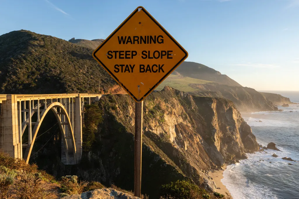 A warning sign in front of Bixby Bridge and Big Sur coastline.