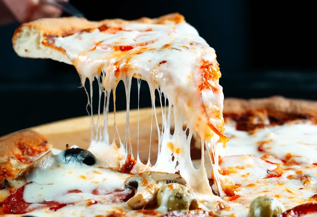 A cheesy pizza slice being pulled away from the rest of the pizza, with strings of melted mozzarella cheese stretching down.