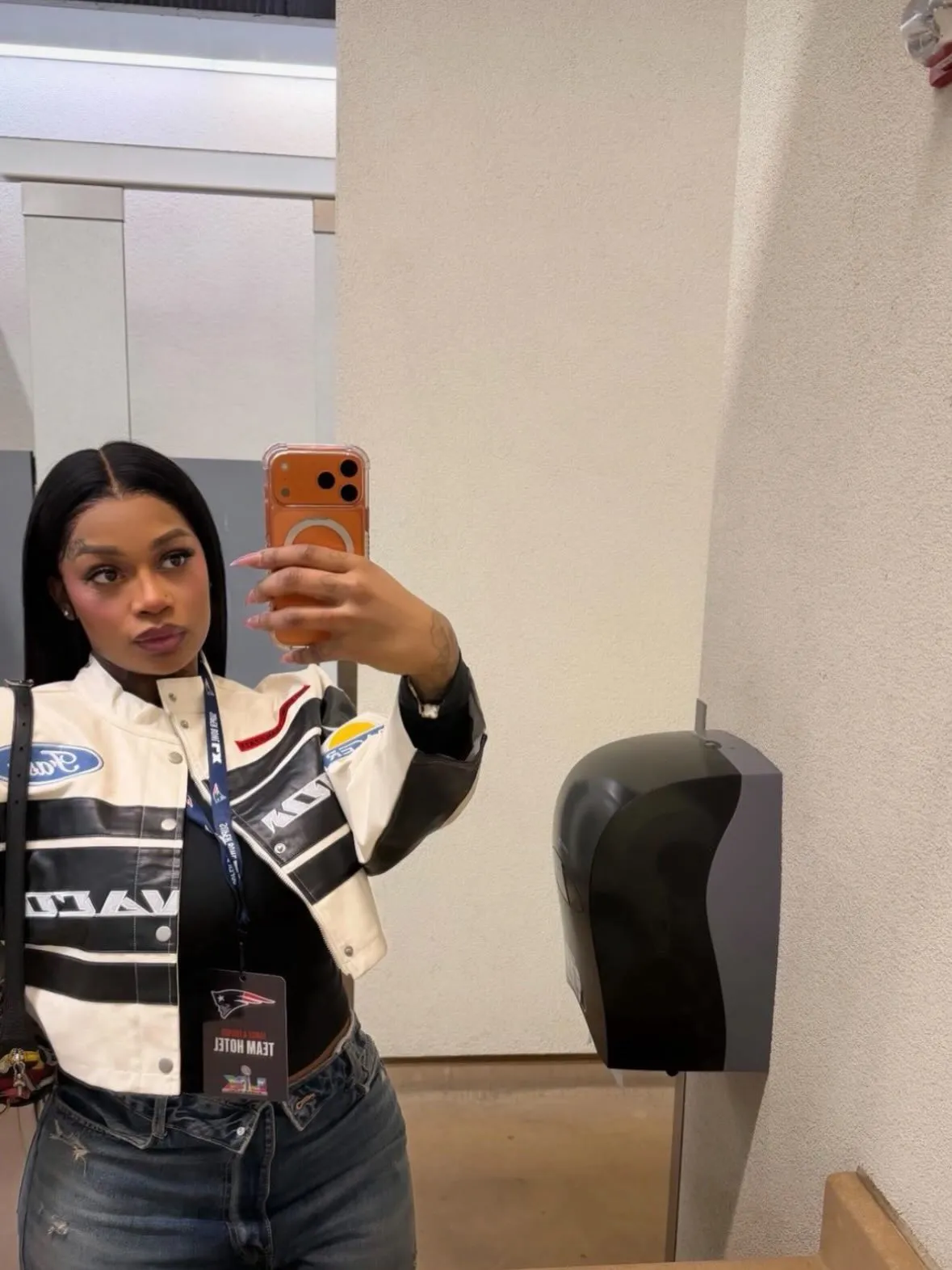 A woman wearing a black and white racing jacket and jeans takes a mirror selfie, showing an all-access pass for the Patriots' team hotel.
