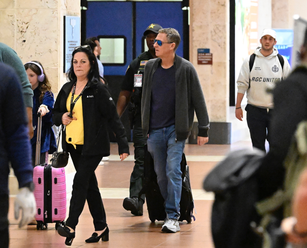 Michael Feldman, Savannah Guthrie’s husband, walking through a Florida airport with a pink suitcase, followed by a child in lavender headphones, and a police officer.