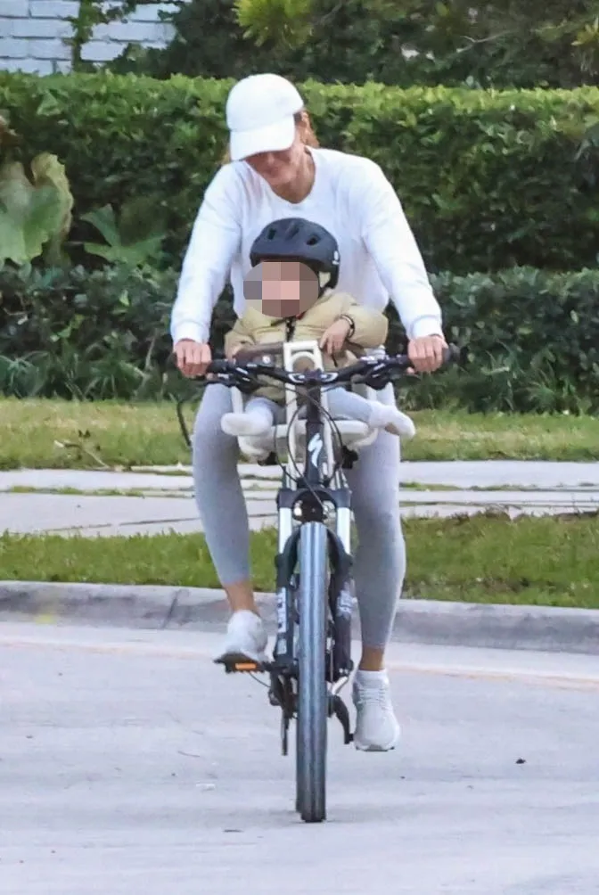 Gisele Bundchen cycling with her one-year-old son, River, in a sunny neighborhood.