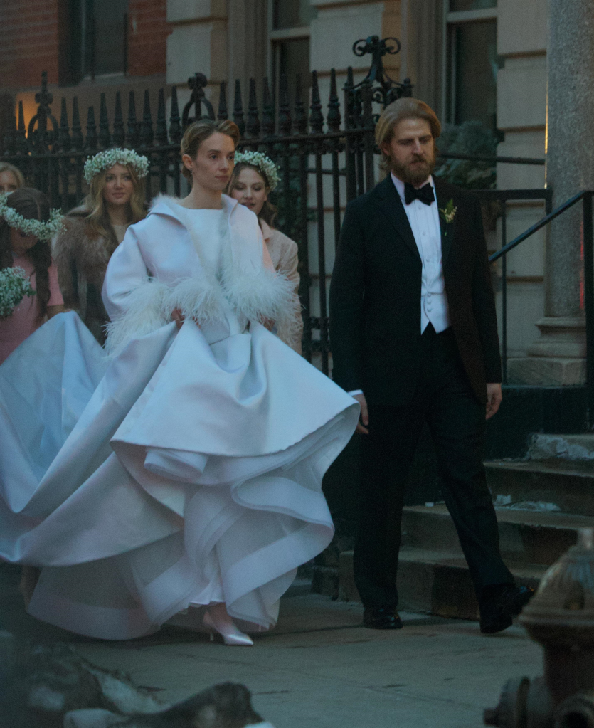 Maya Hawke in a white wedding gown with feathery sleeves and Christian Lee Hutson in a tuxedo, walking outdoors.