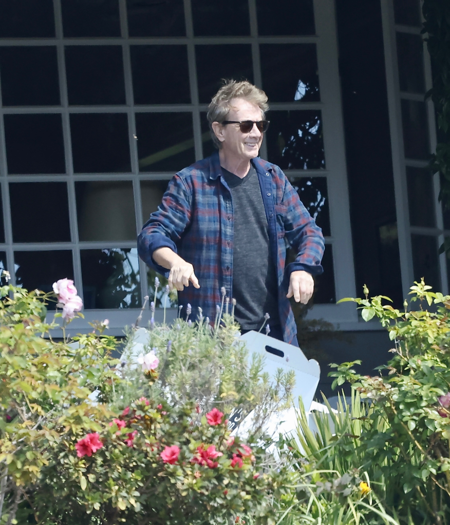 Martin Short smiling outside his home, wearing sunglasses and a plaid shirt, with a window and plants in the background.