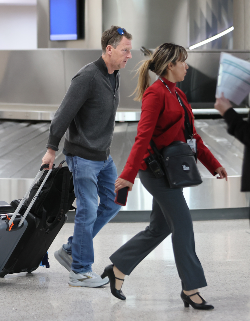 Michael Feldman walking through an airport with his luggage, accompanied by an attendant in a red jacket.