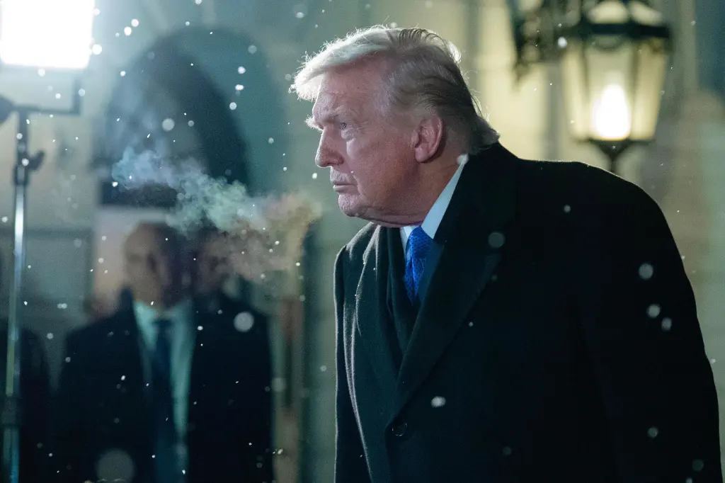 President Donald Trump in a dark overcoat, with visible breath in the cold, walking in what appears to be snowfall.