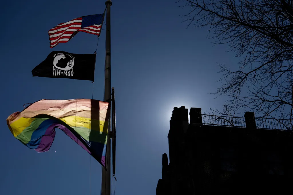 A Pride flag, American flag, and POW-MIA flag fly from a pole against a clear blue sky, with a silhouetted building and tree branches in the background.