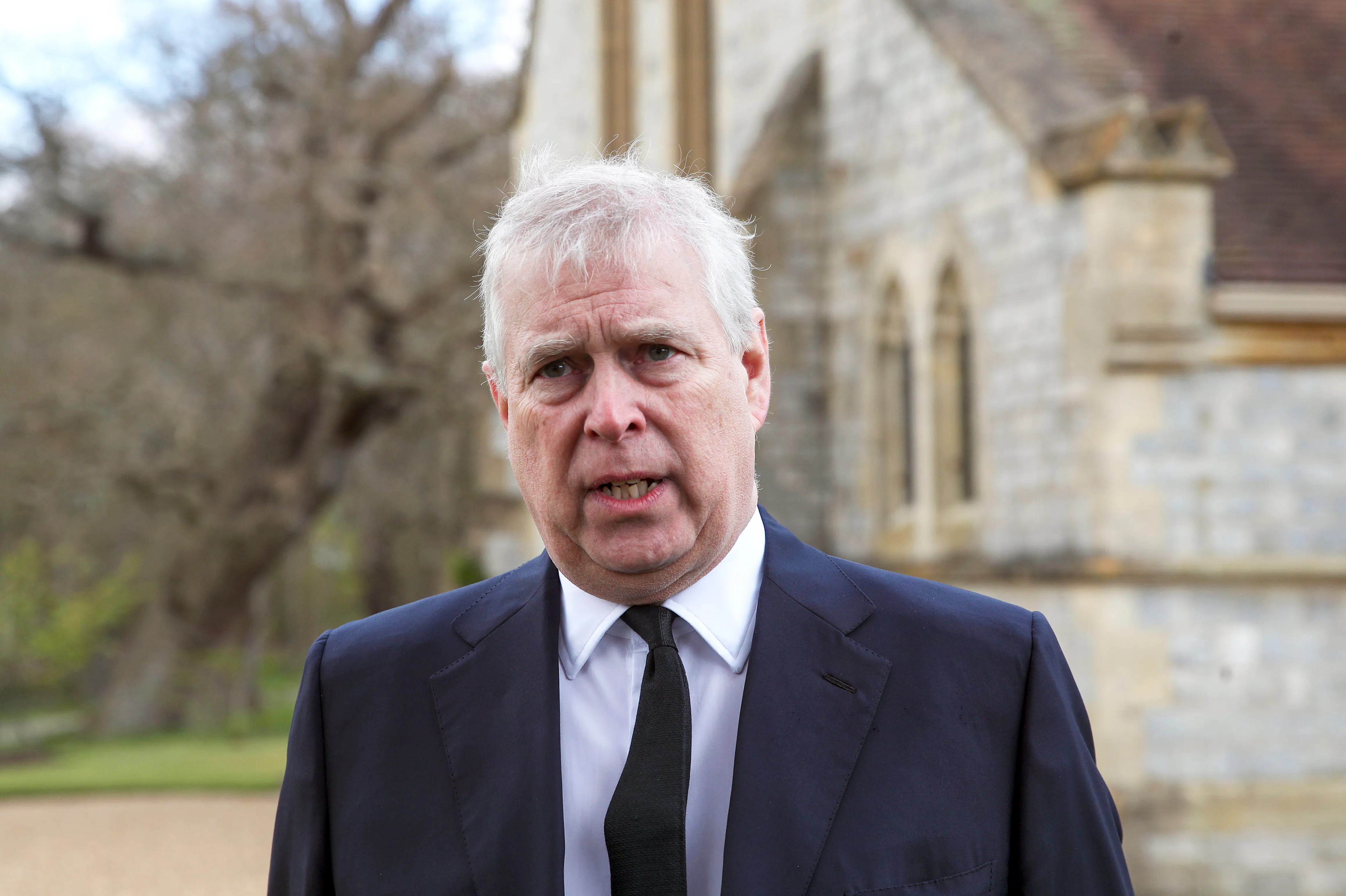 Prince Andrew, Duke of York, wearing a dark suit and black tie, with a white shirt, stands outside a building, mouth slightly open.
