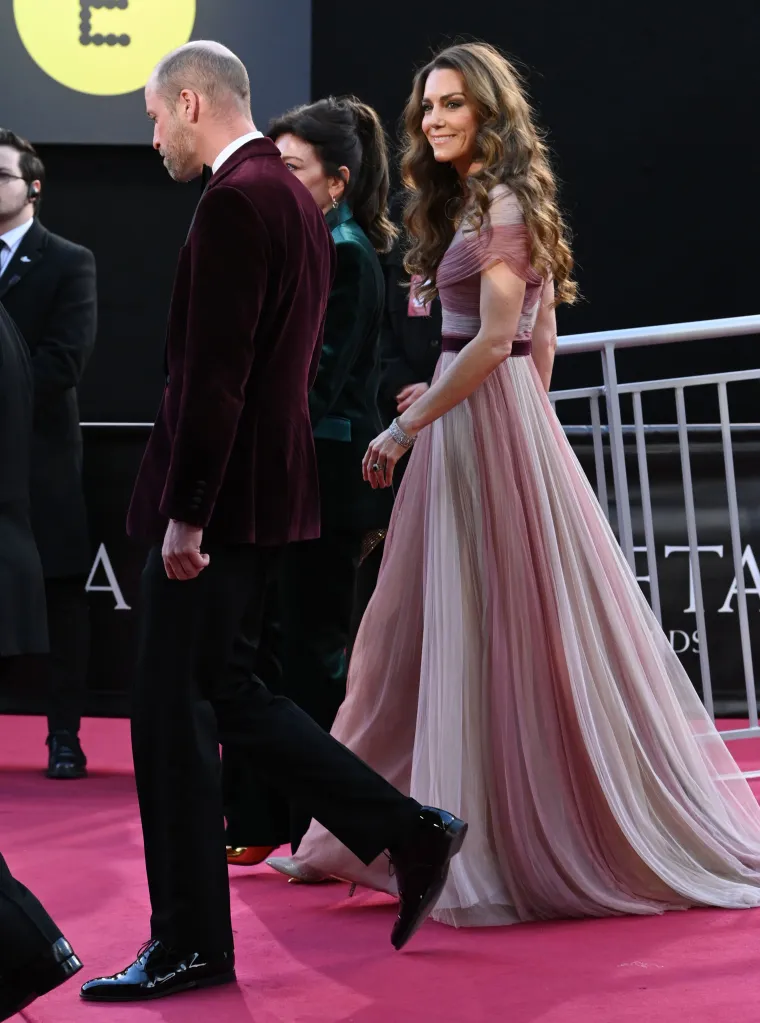 Prince William in a burgundy jacket and Catherine, Princess of Wales, in a pink and white gown, on a red carpet.