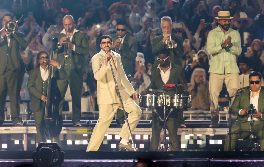 Bad Bunny performs on stage in a light-colored suit, holding a microphone, surrounded by his band members.