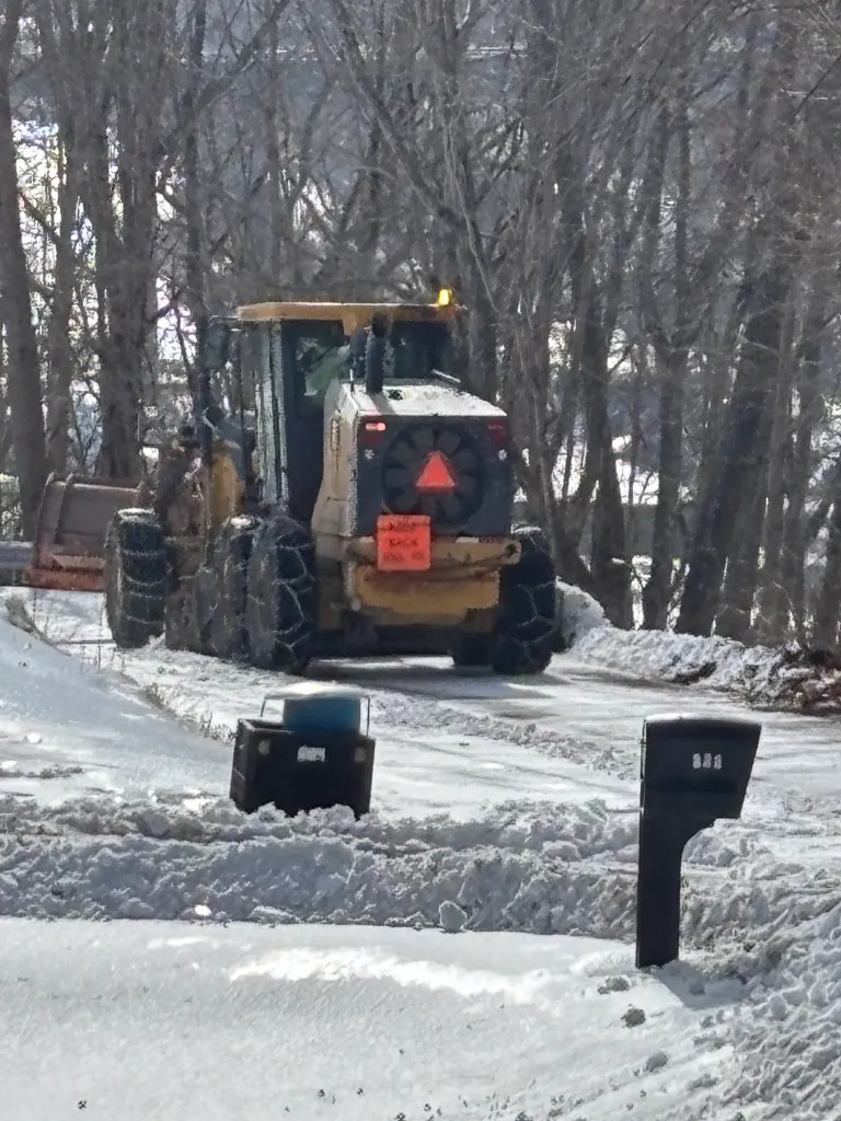 A road grader with tire chains clears snow from a road.