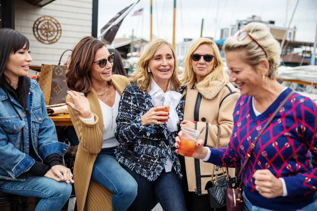 Five women, including Luann de Lesseps, Sonja Morgan, Ramona Singer, and Dorinda Medley, sitting outdoors, smiling and holding drinks.
