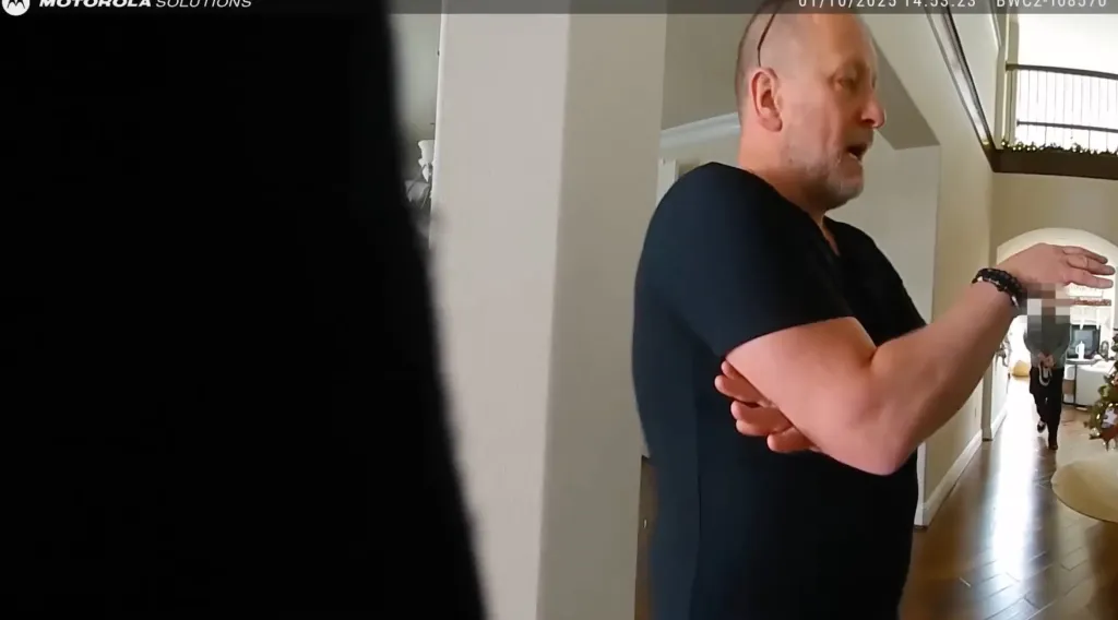 Bodycam footage showing a man with short hair and a black shirt talking, with another person blurred out in the background.