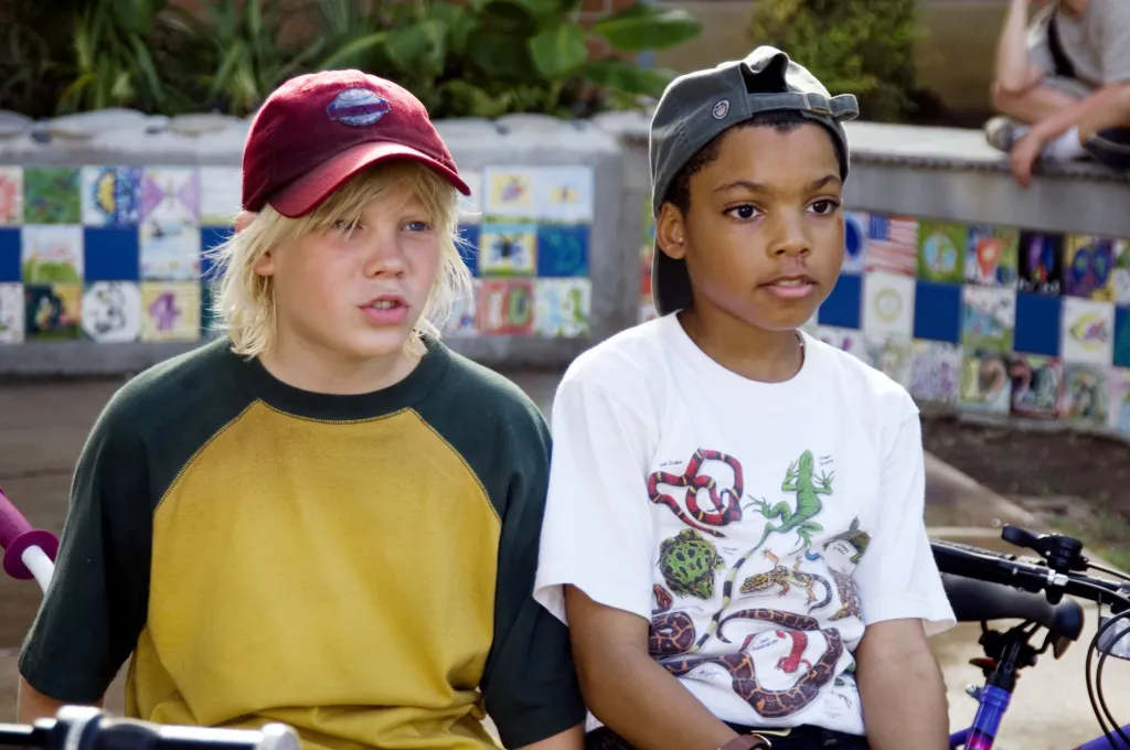 Two boys, one with blonde hair and a red cap, and the other with dark hair and a gray cap, sit together on bicycles.