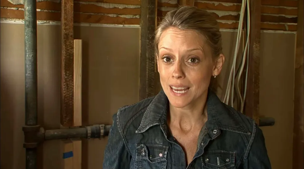 A woman wearing a denim jacket speaks in a room under construction with exposed pipes and wall studs.