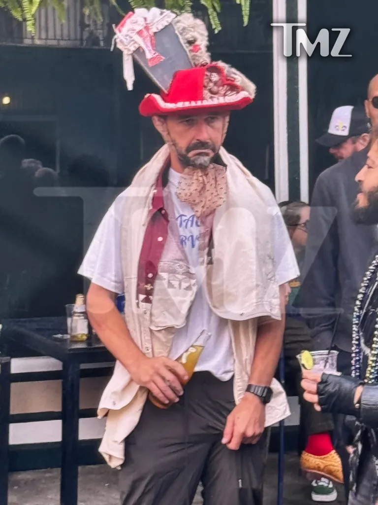 Shia LaBeouf at a Mardi Gras bar crawl, wearing a white t-shirt, a red and white hat, and holding a bottle.