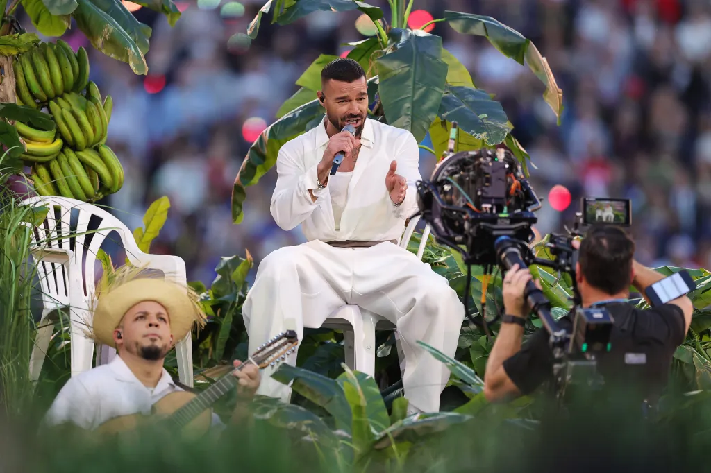 Ricky Martin performing at the Apple Music Super Bowl LX Halftime Show, seated in all white among lush green foliage with a guitarist.