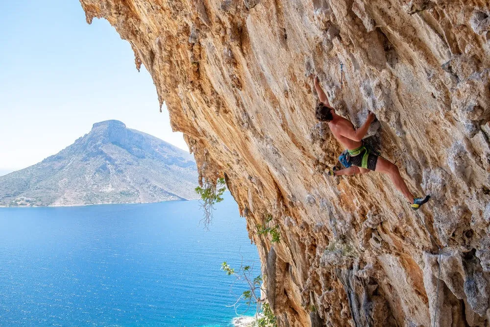 Robert Carroll rock climbing on a cliff face with an ocean and mountain backdrop.