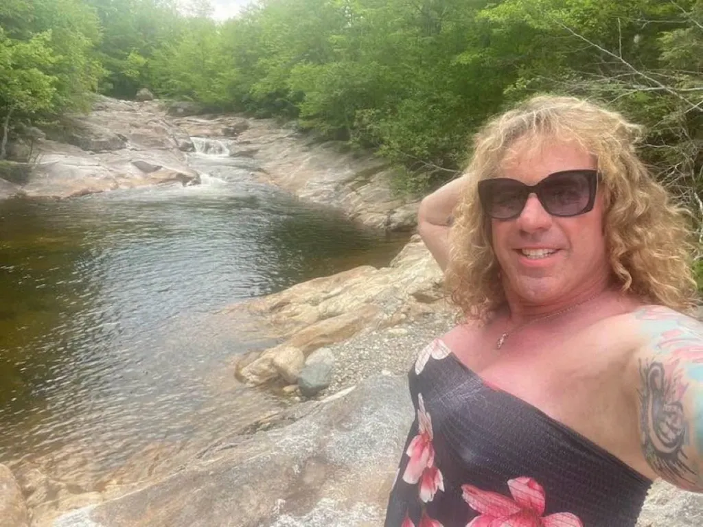 Robert Dorgan, father of a North Providence High School senior, in a black floral dress and sunglasses, posing next to a river.