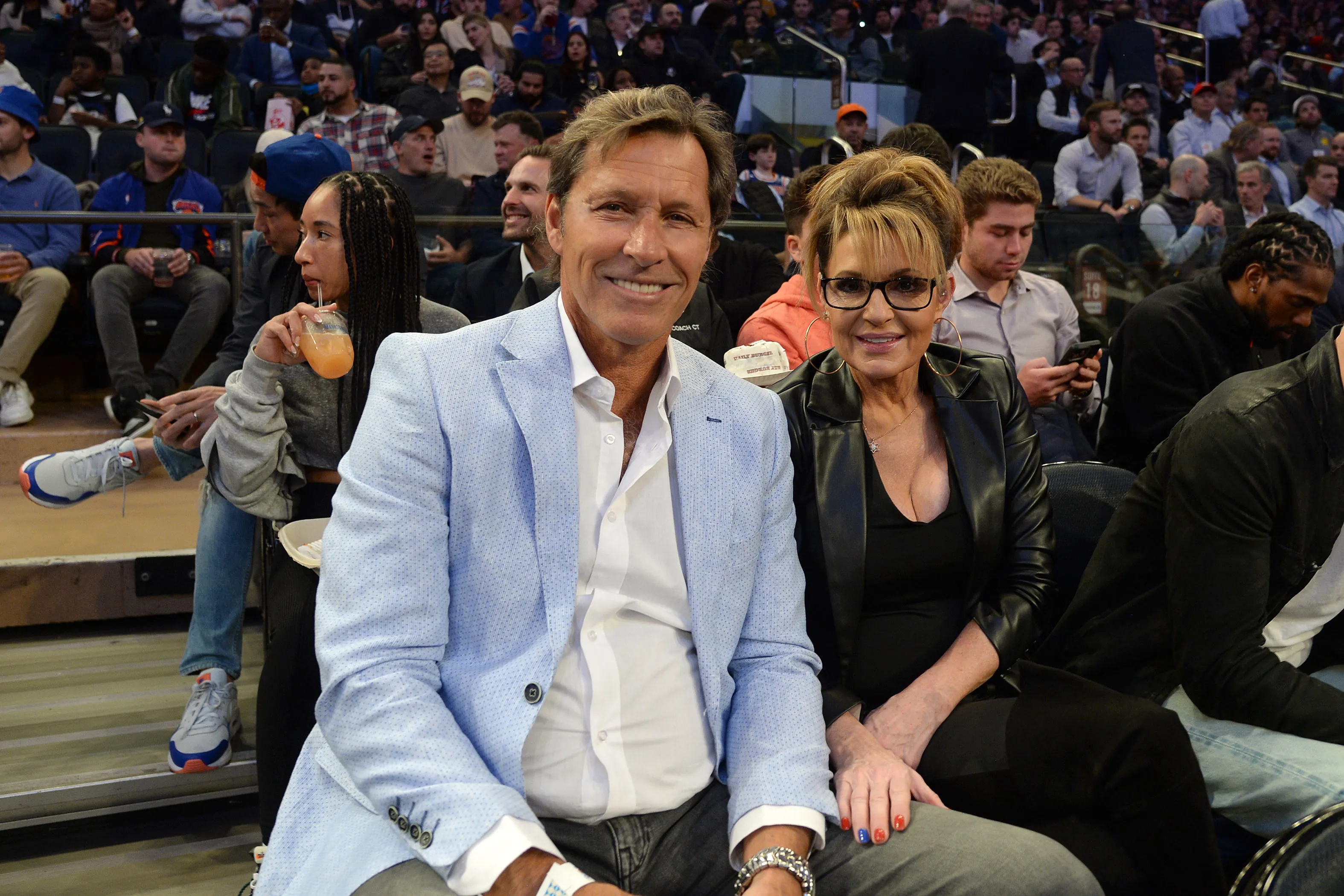 Ron Duguay and Sarah Palin smile while sitting next to each other at a basketball game.
