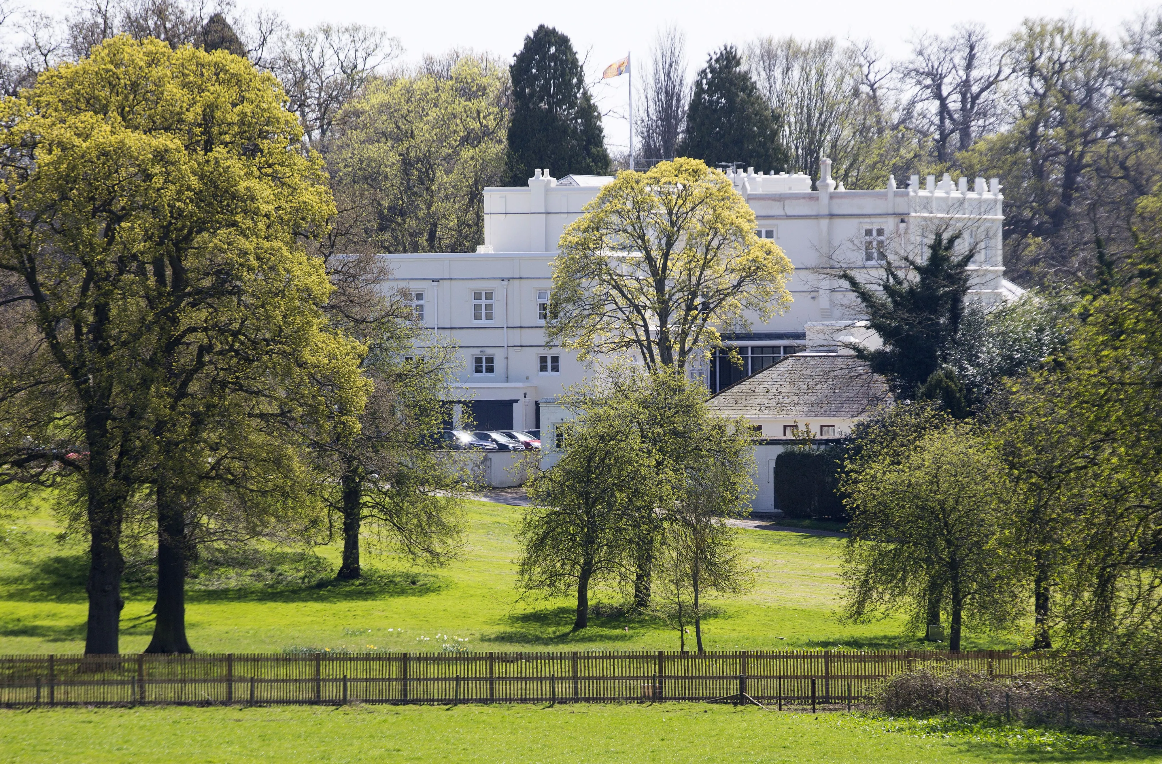 Royal Lodge in Windsor Great Park, the home of The Duke of York, seen from a distance with trees and a wooden fence in the foreground.