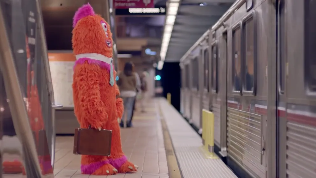 Rude Dude, an orange monster mascot, stands on a subway platform with a briefcase.