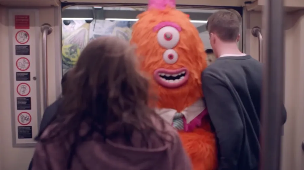 Orange monster mascot with three eyes entering a subway car among people.