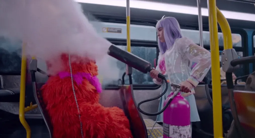 A woman with purple hair spraying a red furry monster on a bus with a fire extinguisher.