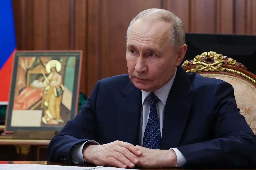Russian President Vladimir Putin in a suit and tie, sitting at a table with his hands clasped, looking slightly to his left.