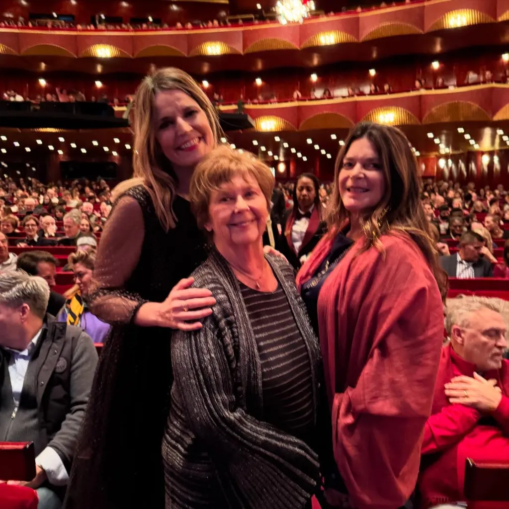Savannah Guthrie,in a black dress and brown top, Nancy Guthrie, in a black and red striped top and matching cardigan, and Annie Guthrie in a red wrap and black top, smiling in a crowded auditorium.